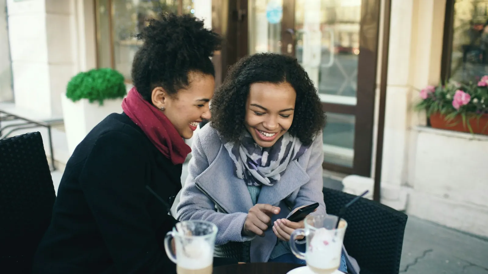 Zwei Frauen lachen in einem Straßencafé über ein Handy